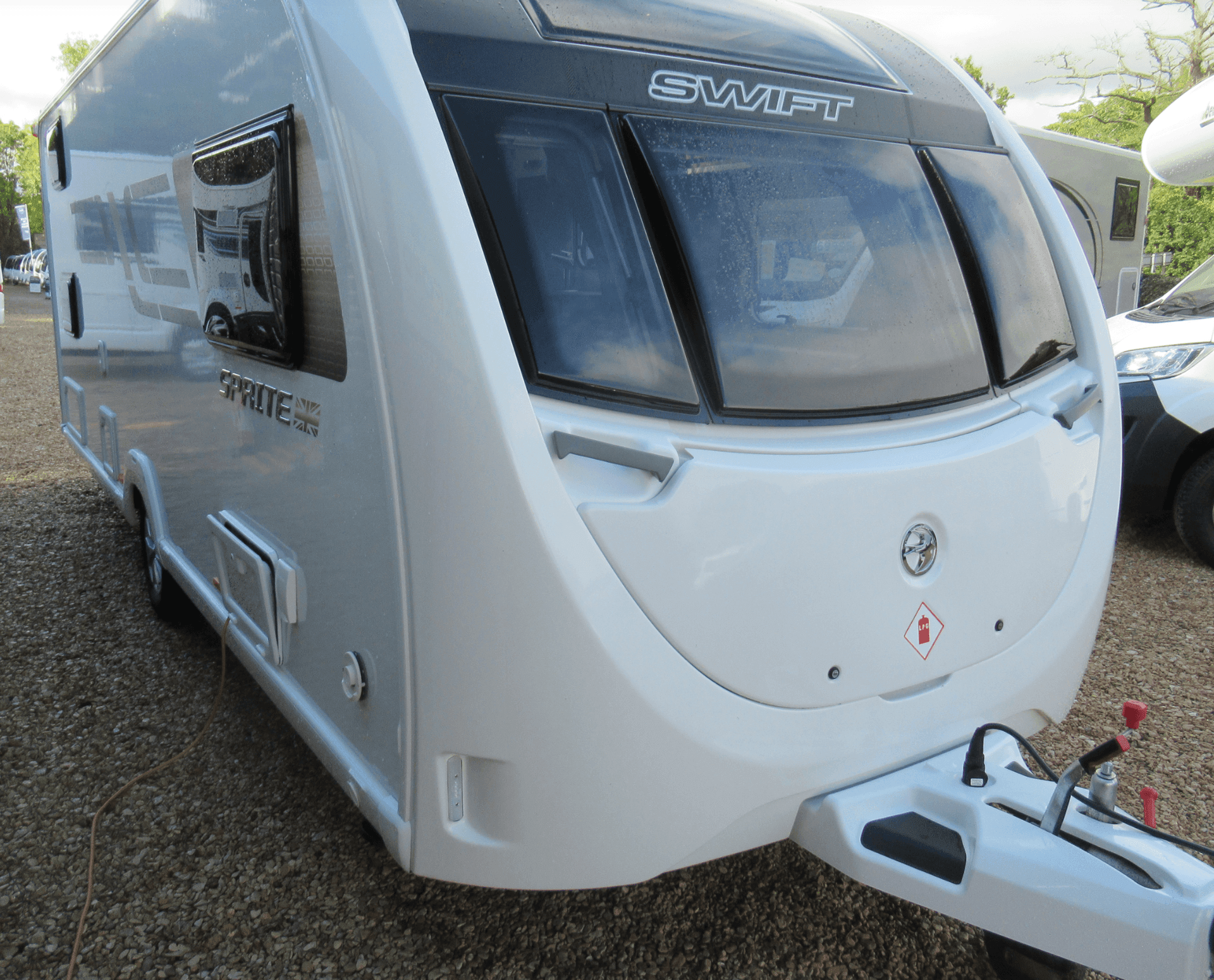 A white Swift Sprite Major 4FB caravan is parked on a gravel surface. It has a streamlined front with large windows and a tow hitch